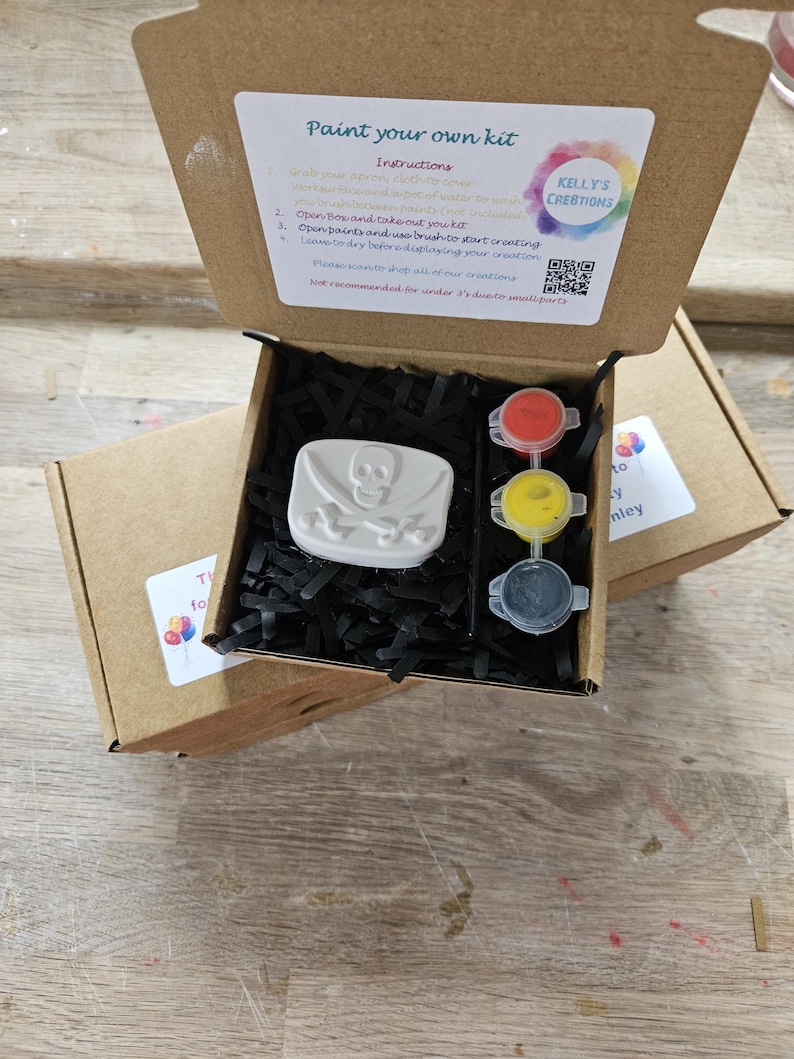 May include: A brown cardboard box containing a paint your own kit with a white skull and crossbones plaster figure, three small paint pots in red, yellow, and black, and a black paintbrush nestled in black paper shred filler material.