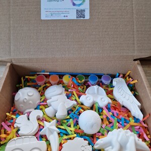 May include: A box filled with colorful paper shreds and white ceramic figurines, including a squirrel, a star, a car, a hedgehog, a dinosaur, a parrot, a mushroom, a dinosaur, a car, a ball, and a paintbrush. The box also includes six small bottles of paint.