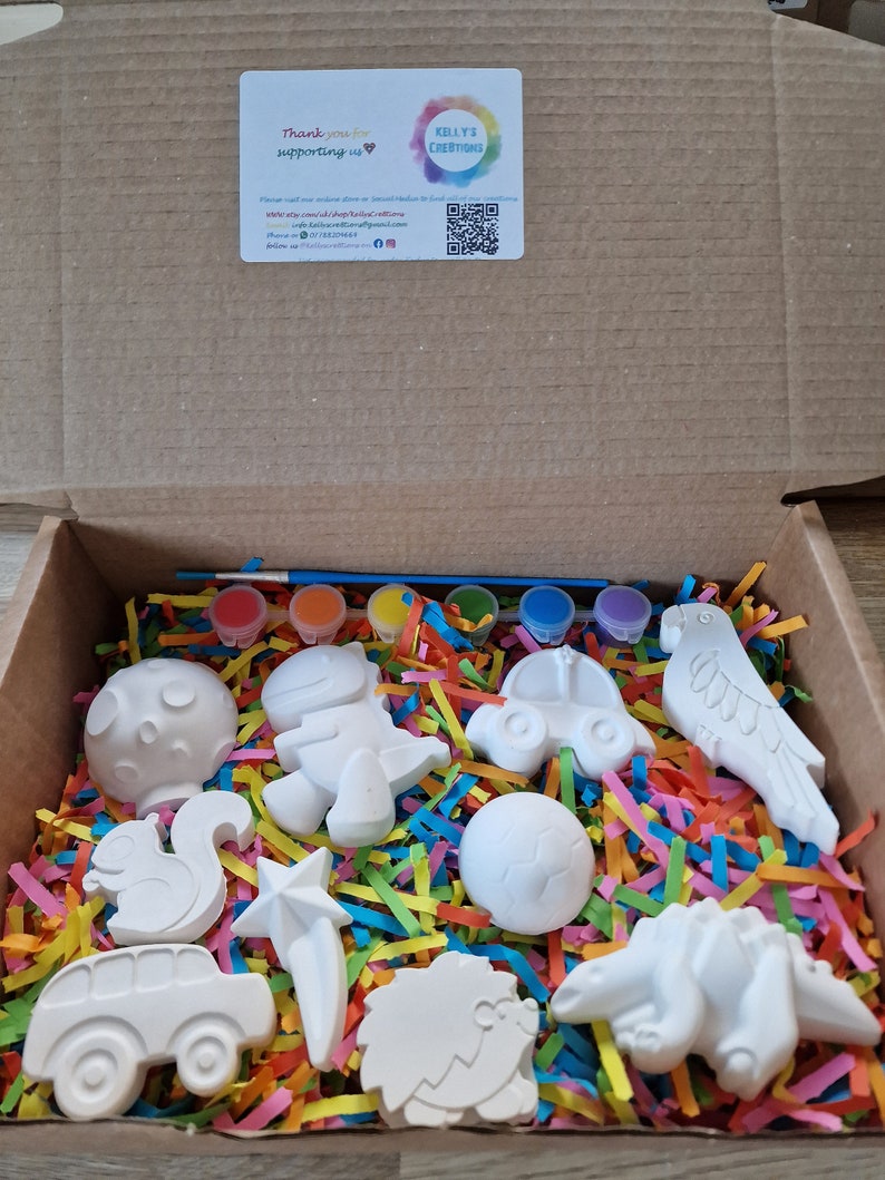 May include: A box filled with colorful paper shreds and white ceramic figurines, including a squirrel, a star, a car, a hedgehog, a dinosaur, a parrot, a mushroom, a dinosaur, a car, a ball, and a paintbrush. The box also includes six small bottles of paint.