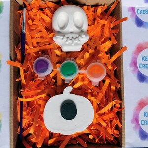 May include: A Halloween-themed craft kit in a cardboard box, featuring a white skull and pumpkin, paint pots in purple, green, and orange, and a paintbrush. The box is filled with orange paper shreds. The text "Kelly's Creations" is visible.