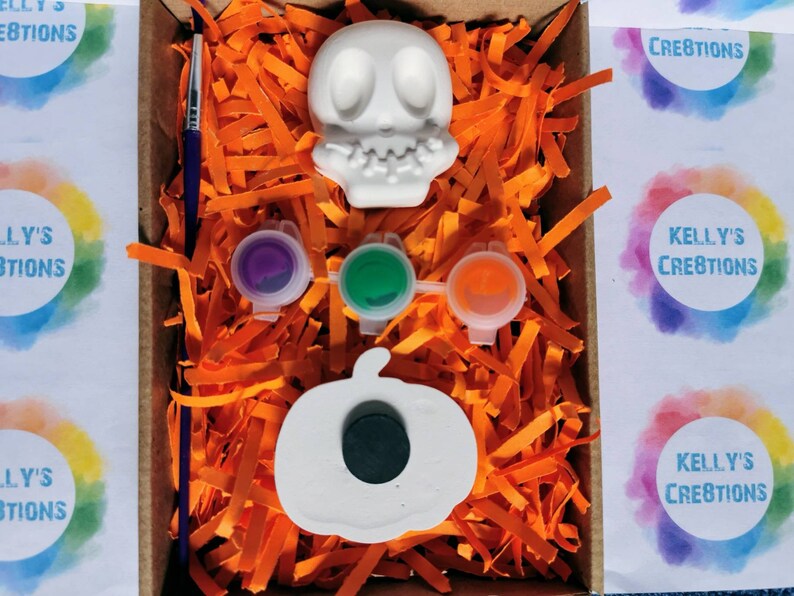 May include: A Halloween-themed craft kit in a cardboard box, featuring a white skull and pumpkin, paint pots in purple, green, and orange, and a paintbrush. The box is filled with orange paper shreds. The text "Kelly's Creations" is visible.