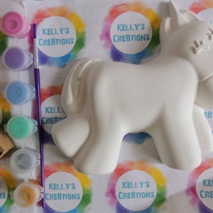 May include: A white ceramic unicorn figurine with paint pots and a paintbrush. The paint pots are pink, purple, blue, green, and yellow. The paint pots have the text "Kelly's Creations" on them.