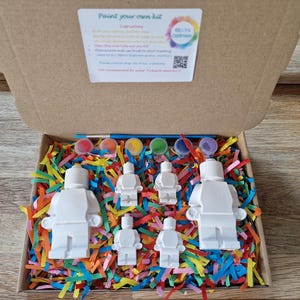 May include: A paint your own kit with five white figurines of people, paint pots, and a paintbrush in a box with colorful shredded paper. The box lid has instructions and the text "Paint your own kit".
