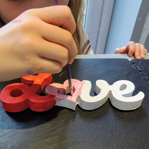May include: A person is painting a wooden "LOVE" sign with pink paint. The sign is made of individual letters and has a heart shape in the middle of the "O". The person is using a purple paintbrush and there are multiple paint pots in the background.