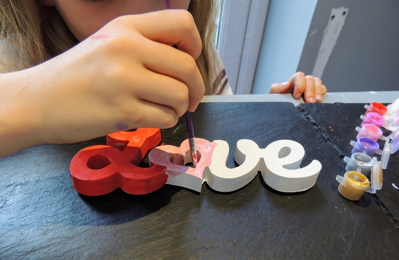 May include: A person is painting a wooden "LOVE" sign with pink paint. The sign is made of individual letters and has a heart shape in the middle of the "O". The person is using a purple paintbrush and there are multiple paint pots in the background.