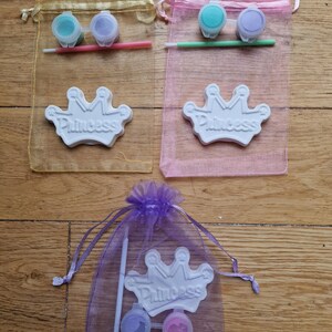 May include: Three organza bags with paint and a ceramic crown for painting. The bags are yellow, pink, and purple. Each bag contains two small paint pots and a paintbrush. The crowns are white with the word "Princess" written on them.