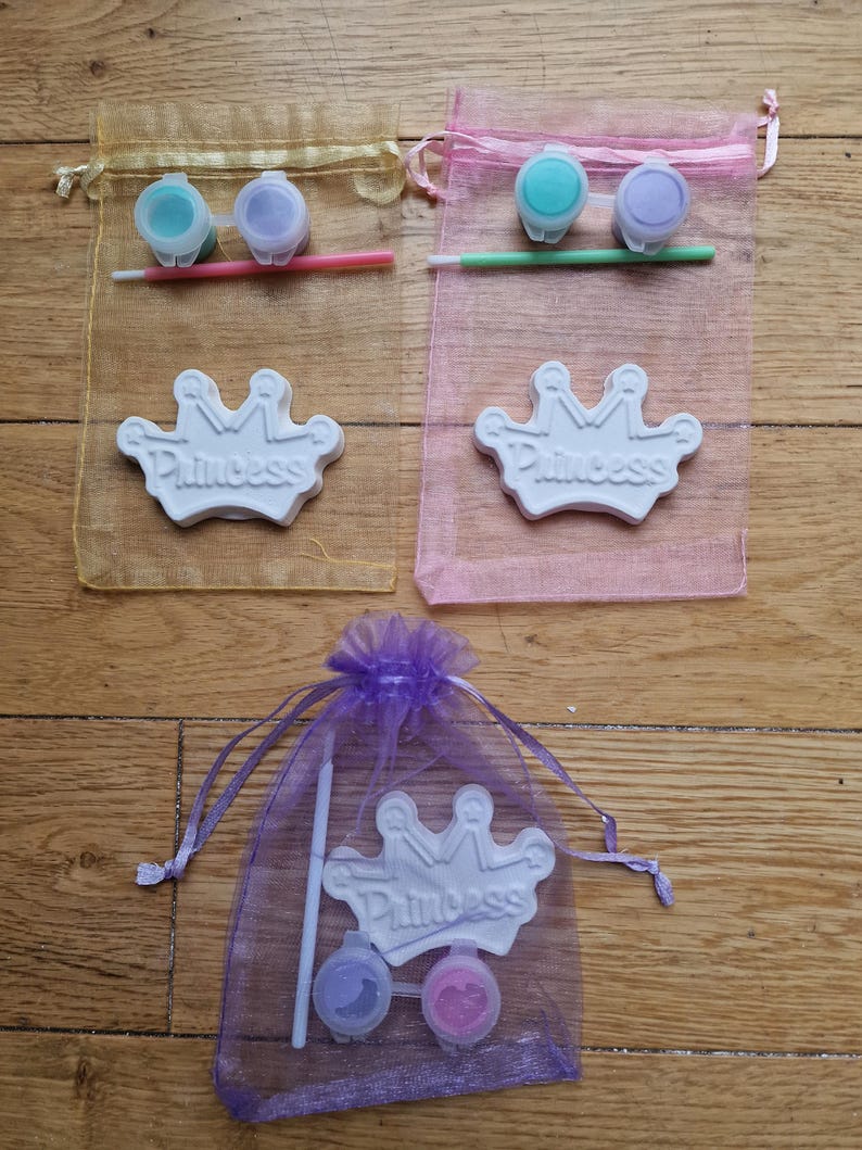 May include: Three organza bags with paint and a ceramic crown for painting. The bags are yellow, pink, and purple. Each bag contains two small paint pots and a paintbrush. The crowns are white with the word "Princess" written on them.