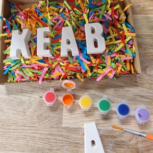 Op de afbeelding: Een set witte keramische letters die de naam "KEAB" spellen in een doos gevuld met kleurrijk versnipperd papier. De letters zijn klaar om te worden geschilderd met de meegeleverde set van zes verfpotjes in rood, oranje, geel, groen, blauw en paars. Een kwast en een enkele letter "A" zijn ook inbegrepen.