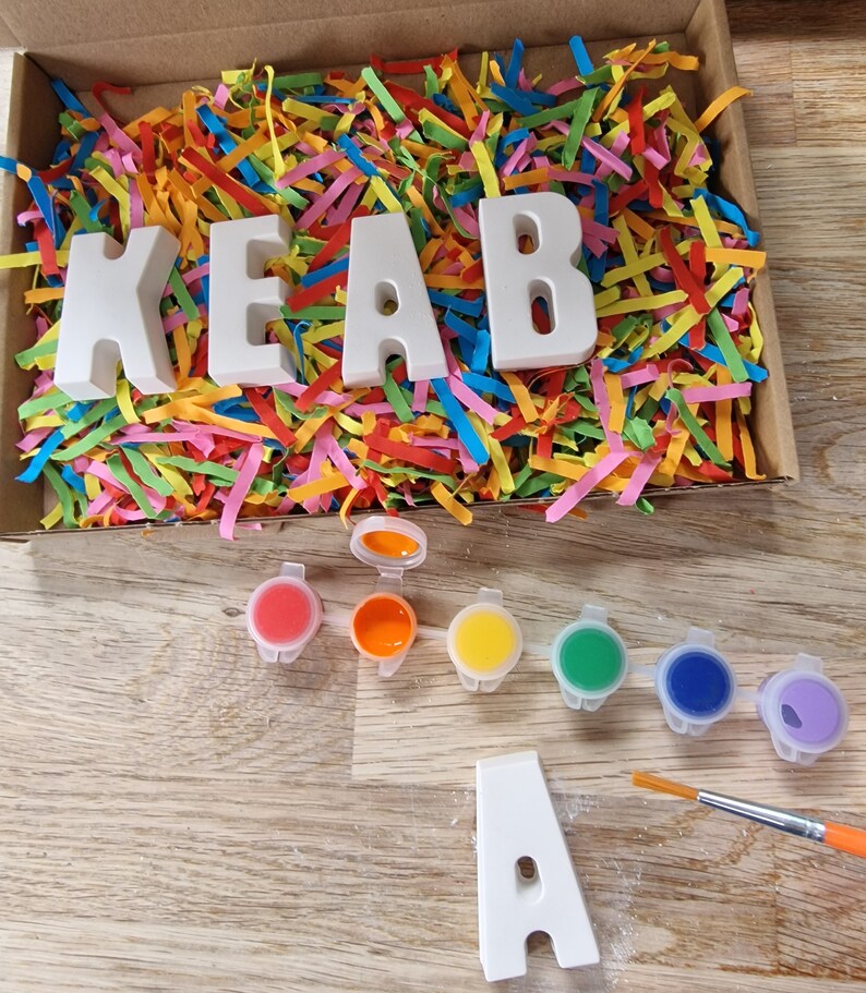 Op de afbeelding: Een set witte keramische letters die de naam "KEAB" spellen in een doos gevuld met kleurrijk versnipperd papier. De letters zijn klaar om te worden geschilderd met de meegeleverde set van zes verfpotjes in rood, oranje, geel, groen, blauw en paars. Een kwast en een enkele letter "A" zijn ook inbegrepen.