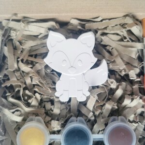 May include: A white ceramic fox figurine with a paint-by-numbers design. The kit includes three small pots of paint: yellow, black, and brown. A paintbrush is also included.