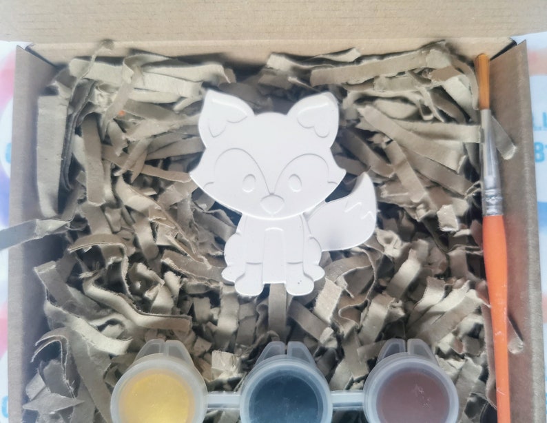 May include: A white ceramic fox figurine with a paint-by-numbers design. The kit includes three small pots of paint: yellow, black, and brown. A paintbrush is also included.