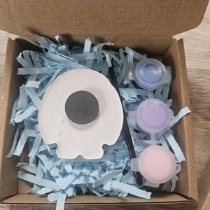 以下が含まれることがあります： 段ボール箱に入ったクラフトキット。白いドクロ型のマグネット、青、紫、ピンクの絵の具が入った3つのペイントポット、筆が特徴です。箱は水色のシュレッダー紙で満たされています。