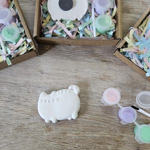 May include: A DIY craft kit featuring a white plaster cat-shaped figure, a small paintbrush, and three small pots of pastel paint in pink, purple, and green. The kit is packaged in a brown cardboard box filled with colourful shredded paper filler.