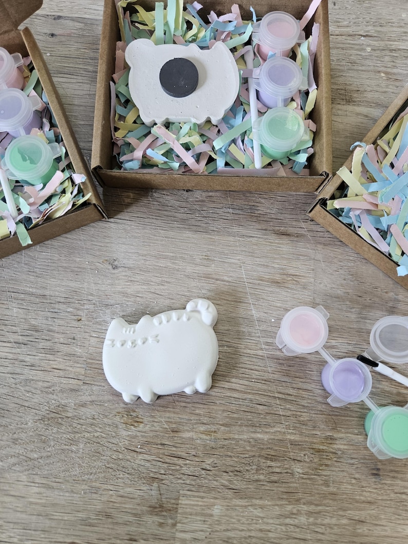 May include: A DIY craft kit featuring a white plaster cat-shaped figure, a small paintbrush, and three small pots of pastel paint in pink, purple, and green. The kit is packaged in a brown cardboard box filled with colourful shredded paper filler.