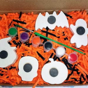 May include: A Halloween-themed craft kit featuring five white wooden shapes: a bat, a ghost, a pumpkin, a skull, and a witch's hat. The shapes have black magnet circles in the centre. The kit also includes a paintbrush, a green paint stick, and six small pots of paint in various colours.