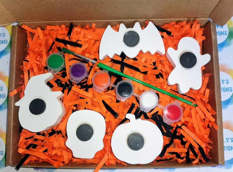 May include: A Halloween-themed craft kit featuring five white wooden shapes: a bat, a ghost, a pumpkin, a skull, and a witch's hat. The shapes have black magnet circles in the centre. The kit also includes a paintbrush, a green paint stick, and six small pots of paint in various colours.