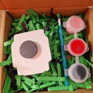 May include: A craft kit in a cardboard box, featuring a white ceramic sheep magnet with a black magnet attached, three paint pots with pink, red, and black paint, a blue paintbrush, and green and black paper shreds. The text "Not recommended for under 3" is visible.