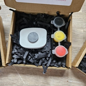 May include: A brown cardboard craft kit box containing a white rectangular object with a circular black magnet on top, three small paint pots in black, yellow, and red, and black paper filler. The box lid features text and a QR code.
