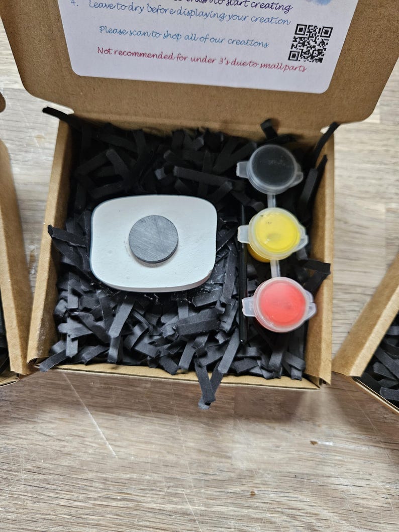 May include: A brown cardboard craft kit box containing a white rectangular object with a circular black magnet on top, three small paint pots in black, yellow, and red, and black paper filler. The box lid features text and a QR code.