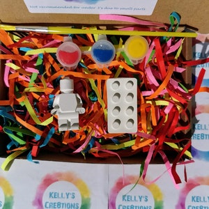 Puede incluir: Una figurita de pl&aacute;stico blanca de una persona, un peque&ntilde;o pincel, tres peque&ntilde;os recipientes de pintura y un molde para hacer peque&ntilde;os ladrillos de pl&aacute;stico. Los art&iacute;culos est&aacute;n en una caja llena de papel triturado de colores. La caja tiene una etiqueta de advertencia que dice "No recomendado para menores de 1 a&ntilde;o debido a piezas peque&ntilde;as".