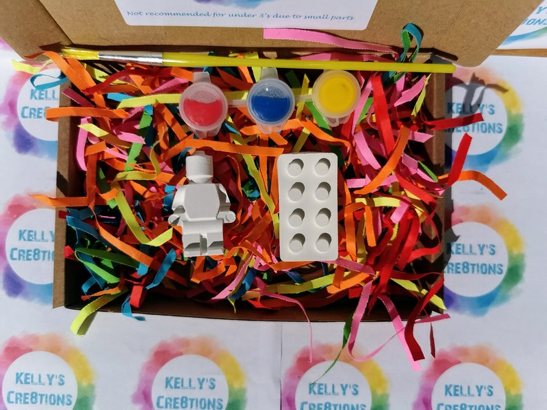 Puede incluir: Una figurita de pl&aacute;stico blanca de una persona, un peque&ntilde;o pincel, tres peque&ntilde;os recipientes de pintura y un molde para hacer peque&ntilde;os ladrillos de pl&aacute;stico. Los art&iacute;culos est&aacute;n en una caja llena de papel triturado de colores. La caja tiene una etiqueta de advertencia que dice "No recomendado para menores de 1 a&ntilde;o debido a piezas peque&ntilde;as".
