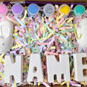 May include: A set of paint-your-own Easter crafts, including a bunny, an egg that says "Happy Easter", and the letters "NAME". The crafts are surrounded by pastel-colored shredded paper and paint pots.