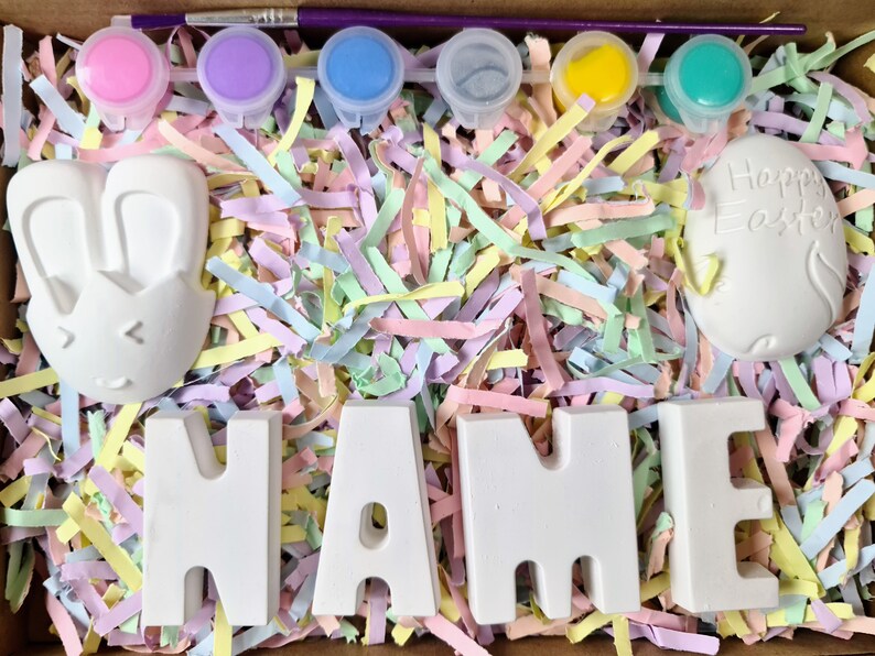May include: A set of paint-your-own Easter crafts, including a bunny, an egg that says "Happy Easter", and the letters "NAME". The crafts are surrounded by pastel-colored shredded paper and paint pots.