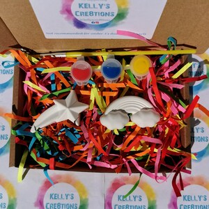 May include: A craft kit with a rainbow and a star shaped ceramic ornament, three small paint pots, a paintbrush, and colorful shredded paper in a cardboard box. The box has the text "Kelly's Creations" on it.