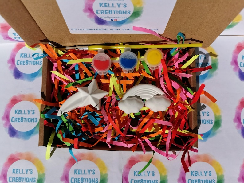 May include: A craft kit with a rainbow and a star shaped ceramic ornament, three small paint pots, a paintbrush, and colorful shredded paper in a cardboard box. The box has the text "Kelly's Creations" on it.