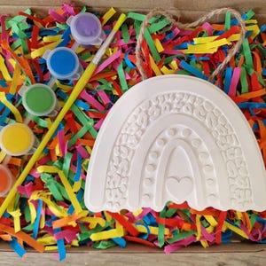 May include: A craft kit with a white rainbow-shaped ceramic piece, paint pots in various colors, and a paintbrush. The kit is nestled in a box filled with colorful shredded paper.