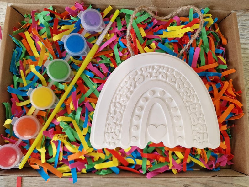 May include: A craft kit with a white rainbow-shaped ceramic piece, paint pots in various colors, and a paintbrush. The kit is nestled in a box filled with colorful shredded paper.