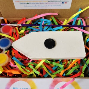 May include: A white wooden rocket ship with a black circle in the center, paint pots, a paintbrush, and colorful paper shreds in a cardboard box. The box has the text "ELLY'S CREATIONS" on it.
