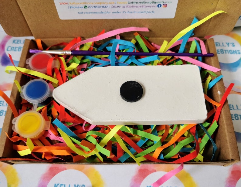 May include: A white wooden rocket ship with a black circle in the center, paint pots, a paintbrush, and colorful paper shreds in a cardboard box. The box has the text "ELLY'S CREATIONS" on it.