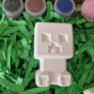 Puede incluir: Una figura blanca inspirada en Minecraft, con un dise&ntilde;o en bloques, sobre tiras de papel verde. Se ven peque&ntilde;os botes de pintura de varios colores, lo que sugiere un proyecto de manualidades.