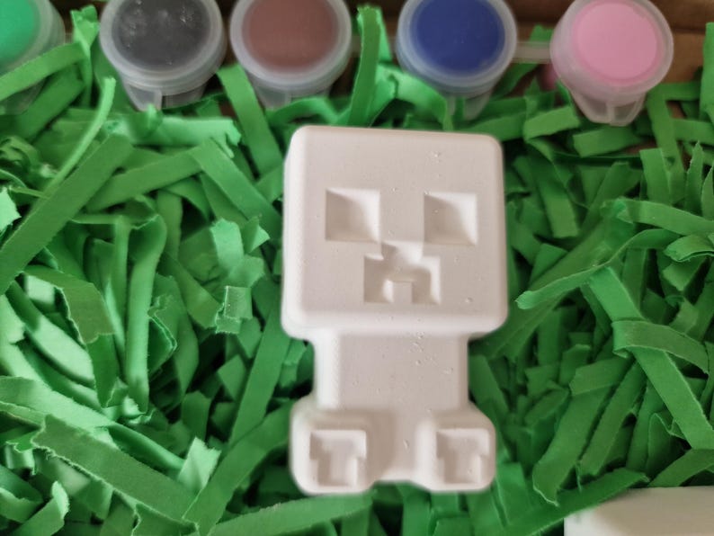Puede incluir: Una figura blanca inspirada en Minecraft, con un dise&ntilde;o en bloques, sobre tiras de papel verde. Se ven peque&ntilde;os botes de pintura de varios colores, lo que sugiere un proyecto de manualidades.
