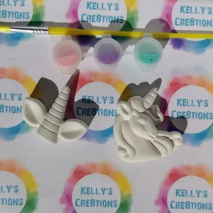 Puede incluir: Un kit de manualidades para pintar unicornios. El kit incluye un pincel, tres botes de pintura con pintura rosa, morada y turquesa, y dos figuras de unicornio blancas. El fondo presenta el texto "KELLY'S CREATIONS" en un c&iacute;rculo arco&iacute;ris.
