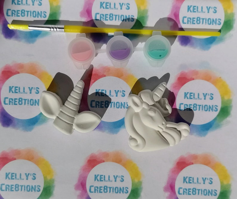 Puede incluir: Un kit de manualidades para pintar unicornios. El kit incluye un pincel, tres botes de pintura con pintura rosa, morada y turquesa, y dos figuras de unicornio blancas. El fondo presenta el texto "KELLY'S CREATIONS" en un c&iacute;rculo arco&iacute;ris.