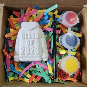 May include: A white ceramic hoodie-shaped ornament with the embossed text "CLASS OF 2026" in a cardboard box filled with colorful paper shreds. The set includes a thin black paintbrush and three small plastic paint pots containing red, blue, and yellow paint.