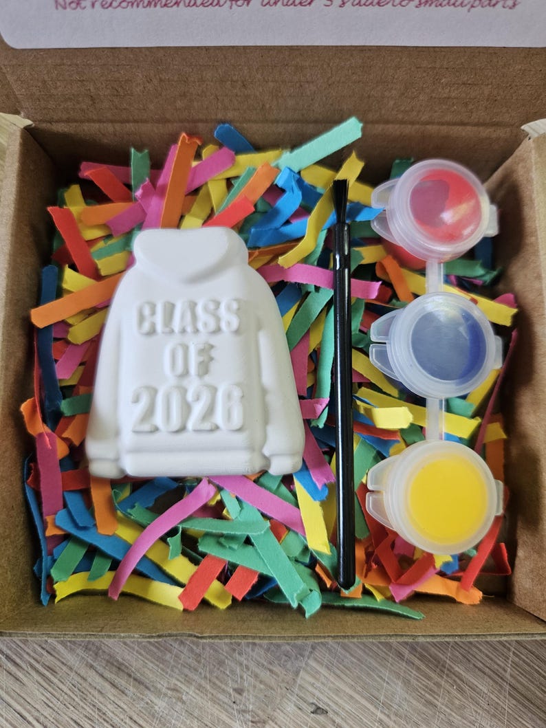 May include: A white ceramic hoodie-shaped ornament with the embossed text "CLASS OF 2026" in a cardboard box filled with colorful paper shreds. The set includes a thin black paintbrush and three small plastic paint pots containing red, blue, and yellow paint.