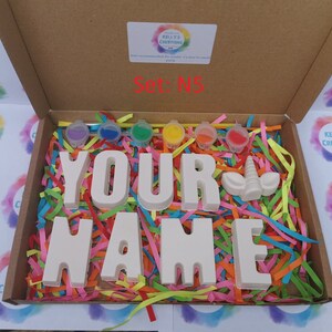 May include: A craft kit in a cardboard box, featuring the words "YOUR NAME" in large, unpainted letters, along with a unicorn horn and paint pots. The box is filled with colorful shredded paper. The text "Set: N5" is visible.