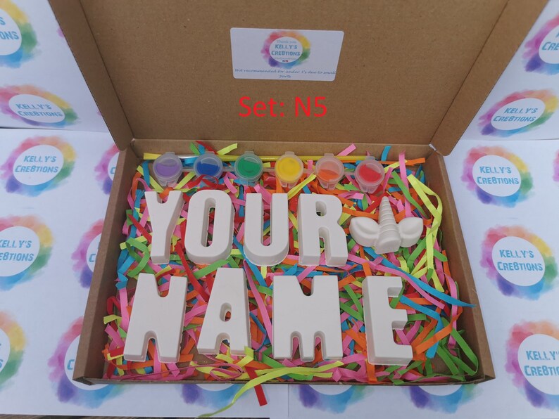 May include: A craft kit in a cardboard box, featuring the words "YOUR NAME" in large, unpainted letters, along with a unicorn horn and paint pots. The box is filled with colorful shredded paper. The text "Set: N5" is visible.