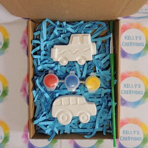 Puede incluir: Un kit de manualidades en una caja de cart&oacute;n, con dos formas de veh&iacute;culos de cer&aacute;mica blanca, botes de pintura roja, azul y amarilla, y un pincel verde. La caja est&aacute; llena de papel triturado azul. El texto "KELLY'S CREATIONS" es visible.