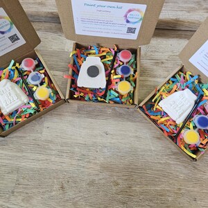 May include: Three brown cardboard boxes containing paint-your-own kits. Each kit includes a white ceramic sweatshirt ornament embossed with "Class of 2026", a small black paintbrush, three circular paint pots in red, blue, and yellow, and colorful paper filler.