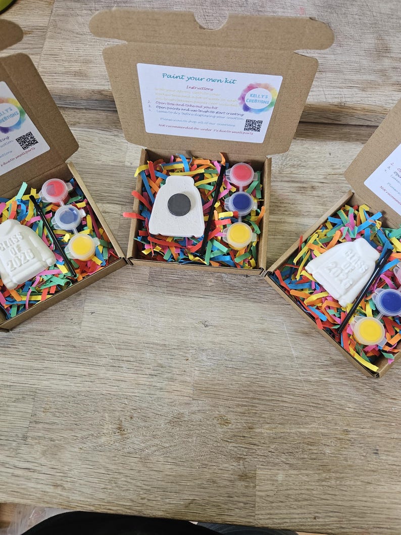 May include: Three brown cardboard boxes containing paint-your-own kits. Each kit includes a white ceramic sweatshirt ornament embossed with "Class of 2026", a small black paintbrush, three circular paint pots in red, blue, and yellow, and colorful paper filler.