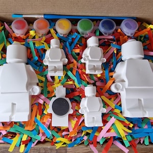 May include: A set of five white ceramic figurines shaped like Lego people, with paint pots and a paintbrush. The figurines are surrounded by colorful shredded paper.