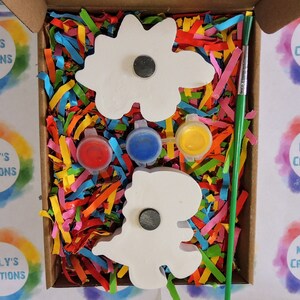 May include: A craft kit with two white wooden dinosaur shapes, three small paint pots in red, blue, and yellow, and a green paintbrush. The kit is packaged in a cardboard box with colorful shredded paper.