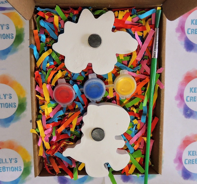 May include: A craft kit with two white wooden dinosaur shapes, three small paint pots in red, blue, and yellow, and a green paintbrush. The kit is packaged in a cardboard box with colorful shredded paper.