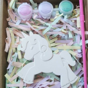 May include: A paint-your-own unicorn kit in a cardboard box. The kit includes a white unicorn figure, three small paint pots in pink, purple, and green, and a pink paintbrush. The box is filled with colorful shredded paper.