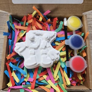 May include: A craft kit in a cardboard box, containing a white plaster figure, colourful paper strips, and paint pots in yellow, blue, and red. The text on the box reads, "Please scan to shop all of our creations. Not recommended for under 3's due to small parts."