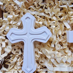 May include: White ceramic cross with a raised design. The cross is sitting on a bed of shredded paper.